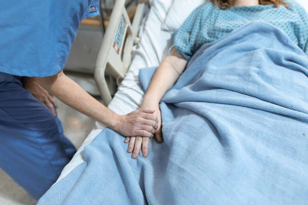 Clinician in blue scrubs holding a patient’s hand in a hospital bed