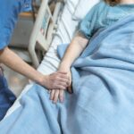 Clinician in blue scrubs holding a patient’s hand in a hospital bed