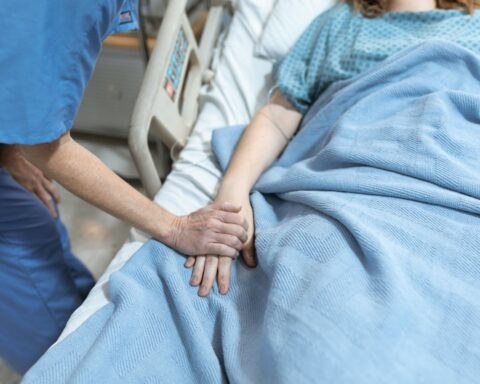 Clinician in blue scrubs holding a patient’s hand in a hospital bed