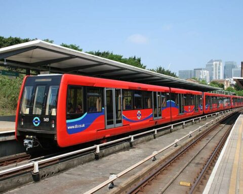 A red and blue Docklands Light Railway train stopped at an outdoor station platform on a sunny day with city buildings in the background.