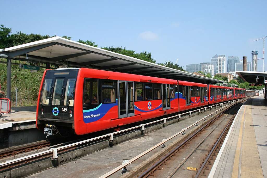 A red and blue Docklands Light Railway train stopped at an outdoor station platform on a sunny day with city buildings in the background.