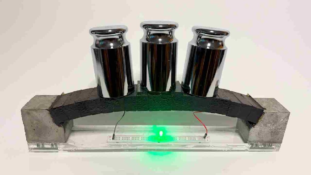 A miniature concrete arch supporting three metal weights, with a green LED light glowing underneath, demonstrating the material's dual functionality for load bearing and powering electronics.