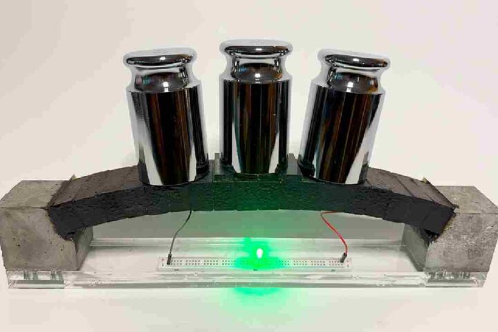 A miniature concrete arch supporting three metal weights, with a green LED light glowing underneath, demonstrating the material's dual functionality for load bearing and powering electronics.