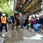 MTA officials and community members, including a person in a wheelchair, participate in the groundbreaking ceremony for the Middletown Road station accessibility project, shoveling dirt to mark the start of construction.