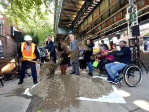 MTA officials and community members, including a person in a wheelchair, participate in the groundbreaking ceremony for the Middletown Road station accessibility project, shoveling dirt to mark the start of construction.