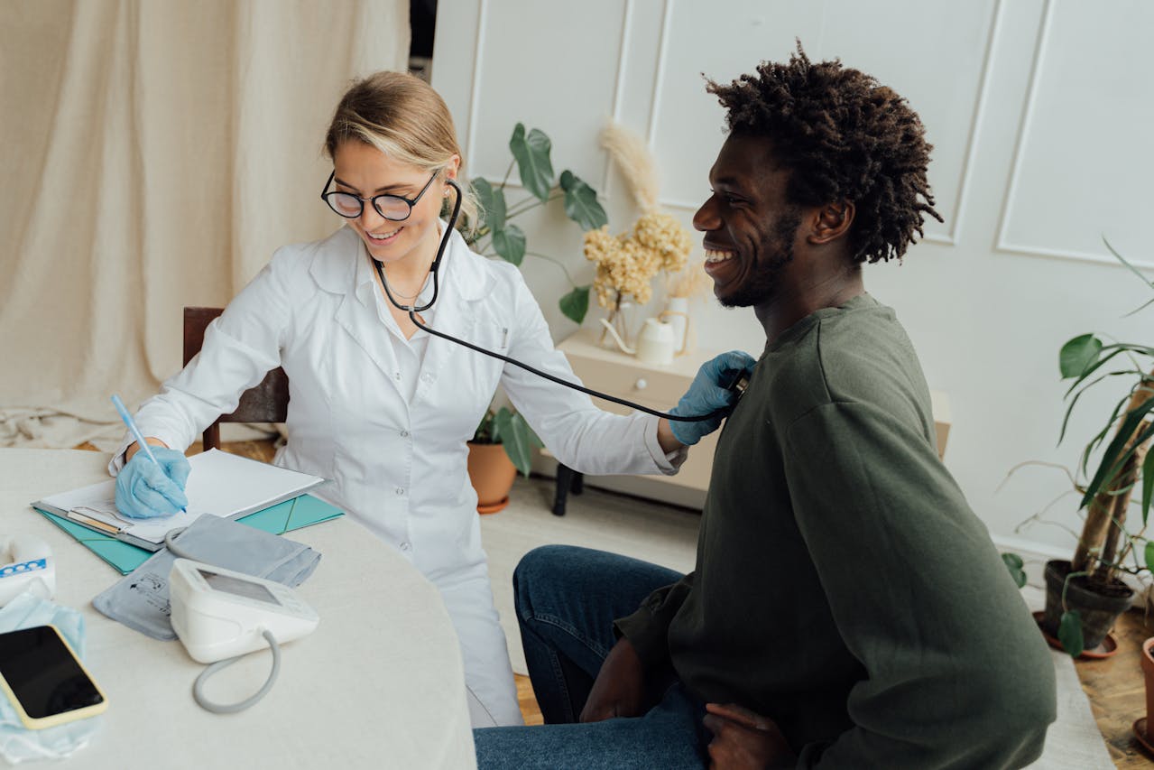 Female doctor in white coat using stethoscope to examine smiling Black male patient during a medical appointment in a bright office setting with plants in the background.