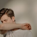 Person demonstrating proper cough etiquette by sneezing or coughing into their elbow rather than their hands, wearing a white shirt against a neutral background.