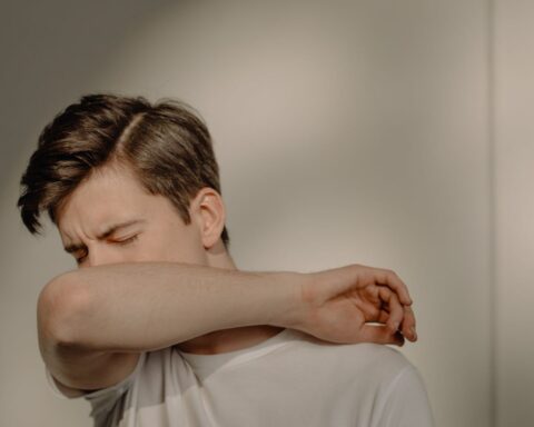 Person demonstrating proper cough etiquette by sneezing or coughing into their elbow rather than their hands, wearing a white shirt against a neutral background.