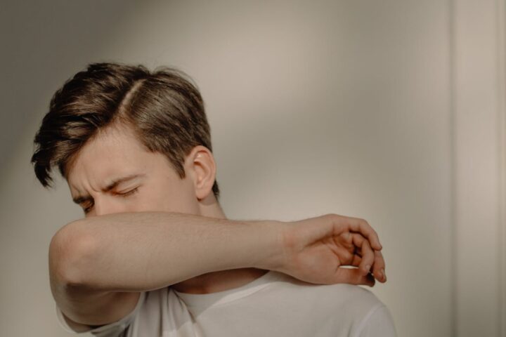 Person demonstrating proper cough etiquette by sneezing or coughing into their elbow rather than their hands, wearing a white shirt against a neutral background.