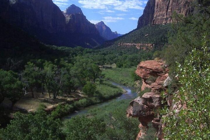 A lush green valley with a winding river and towering red rock cliffs under a dramatic sky at Zion National Park, representing the natural resources at risk during government shutdowns.