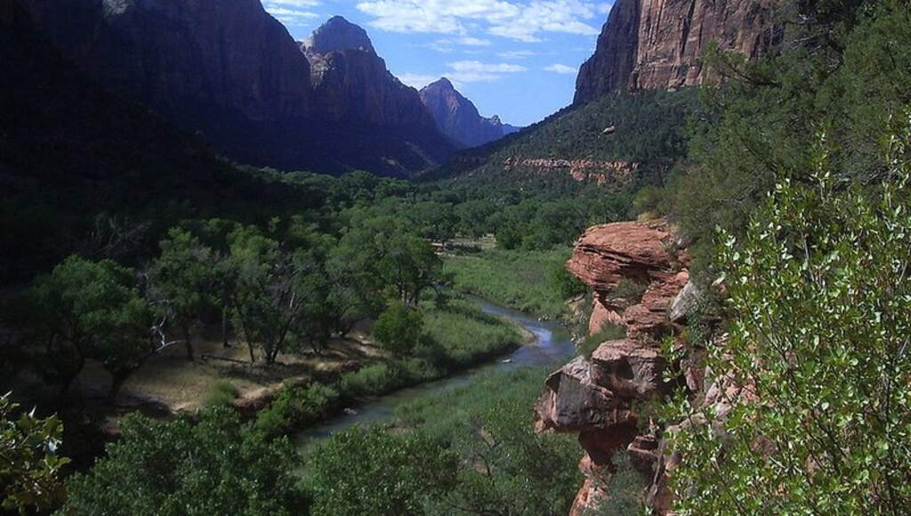 A lush green valley with a winding river and towering red rock cliffs under a dramatic sky at Zion National Park, representing the natural resources at risk during government shutdowns.