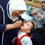 A healthcare worker, wearing a white headscarf and blue tunic, administers oral polio vaccine drops to a young child in Pakistan. The child, dressed in a red and white outfit with a red bow in their hair, has their mouth open. Another woman is visible in the background, out of focus.