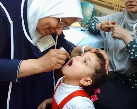 A healthcare worker, wearing a white headscarf and blue tunic, administers oral polio vaccine drops to a young child in Pakistan. The child, dressed in a red and white outfit with a red bow in their hair, has their mouth open. Another woman is visible in the background, out of focus.