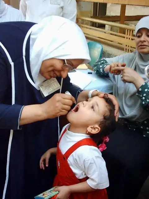 A healthcare worker, wearing a white headscarf and blue tunic, administers oral polio vaccine drops to a young child in Pakistan. The child, dressed in a red and white outfit with a red bow in their hair, has their mouth open. Another woman is visible in the background, out of focus.
