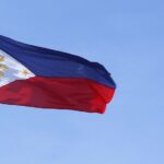 Philippine national flag with a horizontal blue and red bicolor design featuring a white triangle at the hoist containing a golden sun with eight rays and three five-pointed stars against a blue sky background.