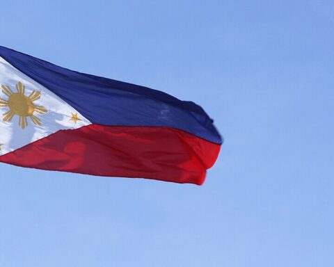 Philippine national flag with a horizontal blue and red bicolor design featuring a white triangle at the hoist containing a golden sun with eight rays and three five-pointed stars against a blue sky background.