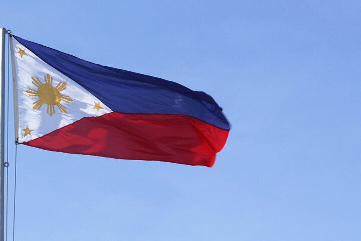 Philippine national flag with a horizontal blue and red bicolor design featuring a white triangle at the hoist containing a golden sun with eight rays and three five-pointed stars against a blue sky background.