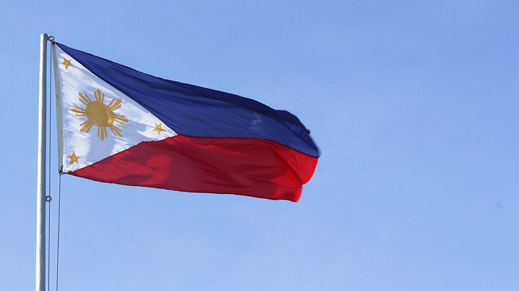 Philippine national flag with a horizontal blue and red bicolor design featuring a white triangle at the hoist containing a golden sun with eight rays and three five-pointed stars against a blue sky background.