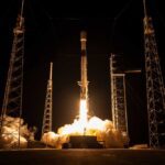 Falcon 9 lifts off at night from SLC-40 for Project Kuiper KF-03, framed by four lightning towers and heavy exhaust plume.