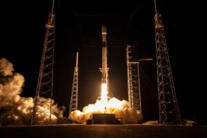 Falcon 9 lifts off at night from SLC-40 for Project Kuiper KF-03, framed by four lightning towers and heavy exhaust plume.