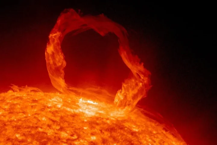 A vibrant red-orange solar flare arc erupting from the sun's surface against the black background of space