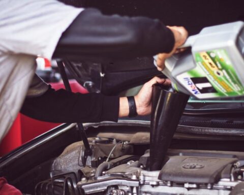 Mechanic wearing work clothes leaning over an open car hood, inspecting the engine in a garage.