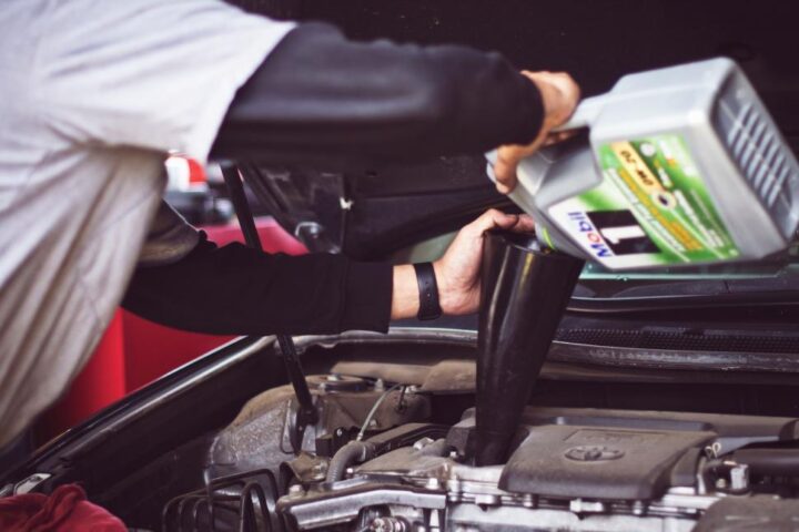 Mechanic wearing work clothes leaning over an open car hood, inspecting the engine in a garage.