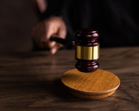 A judge's gavel with wooden base on a dark wooden table with blurred figure in the background, representing legal proceedings and court cases.
