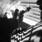 A black and white close-up of a person holding a vaping device, with vapor exhaling from their mouth, highlighting the act of vaping.