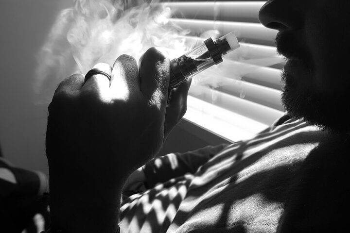 A black and white close-up of a person holding a vaping device, with vapor exhaling from their mouth, highlighting the act of vaping.