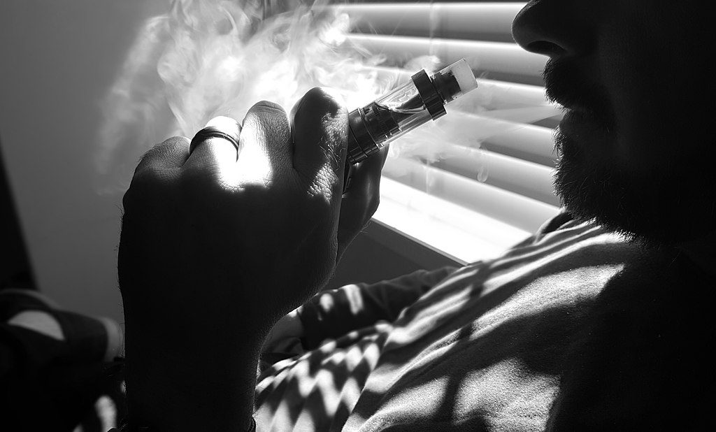 A black and white close-up of a person holding a vaping device, with vapor exhaling from their mouth, highlighting the act of vaping.