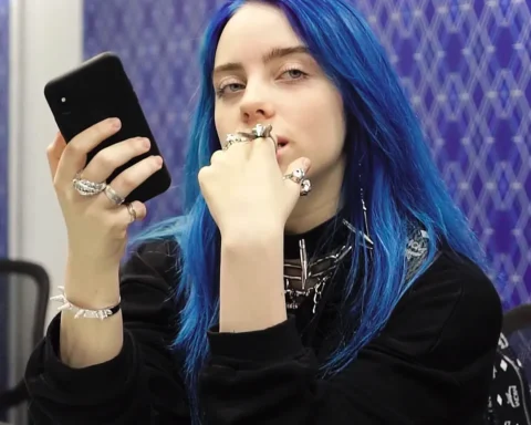 Billie Eilish in a store in 2018, holding a smartphone, blue-tinted hair visible, captured from a YouTube video, used as a feature image for coverage of her $11.5M tour donation to climate justice and food equity.