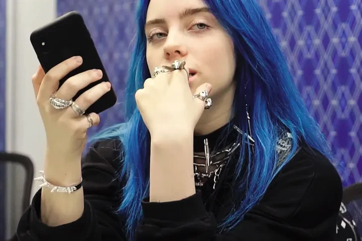 Billie Eilish in a store in 2018, holding a smartphone, blue-tinted hair visible, captured from a YouTube video, used as a feature image for coverage of her $11.5M tour donation to climate justice and food equity.
