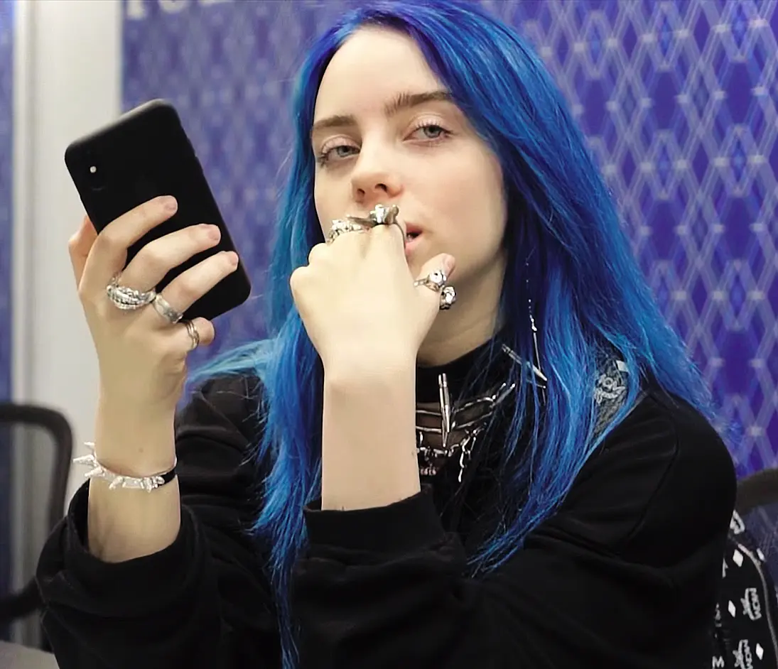 Billie Eilish in a store in 2018, holding a smartphone, blue-tinted hair visible, captured from a YouTube video, used as a feature image for coverage of her $11.5M tour donation to climate justice and food equity.