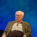 James D. Watson seated on a stage with a blue backdrop, wearing glasses and a jacket, photographed in 2006 during a public talk.