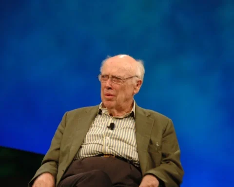 James D. Watson seated on a stage with a blue backdrop, wearing glasses and a jacket, photographed in 2006 during a public talk.