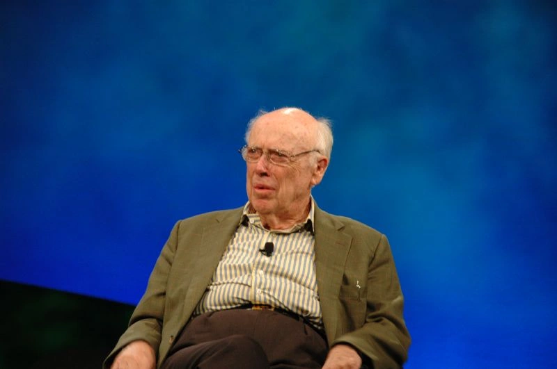 James D. Watson seated on a stage with a blue backdrop, wearing glasses and a jacket, photographed in 2006 during a public talk.