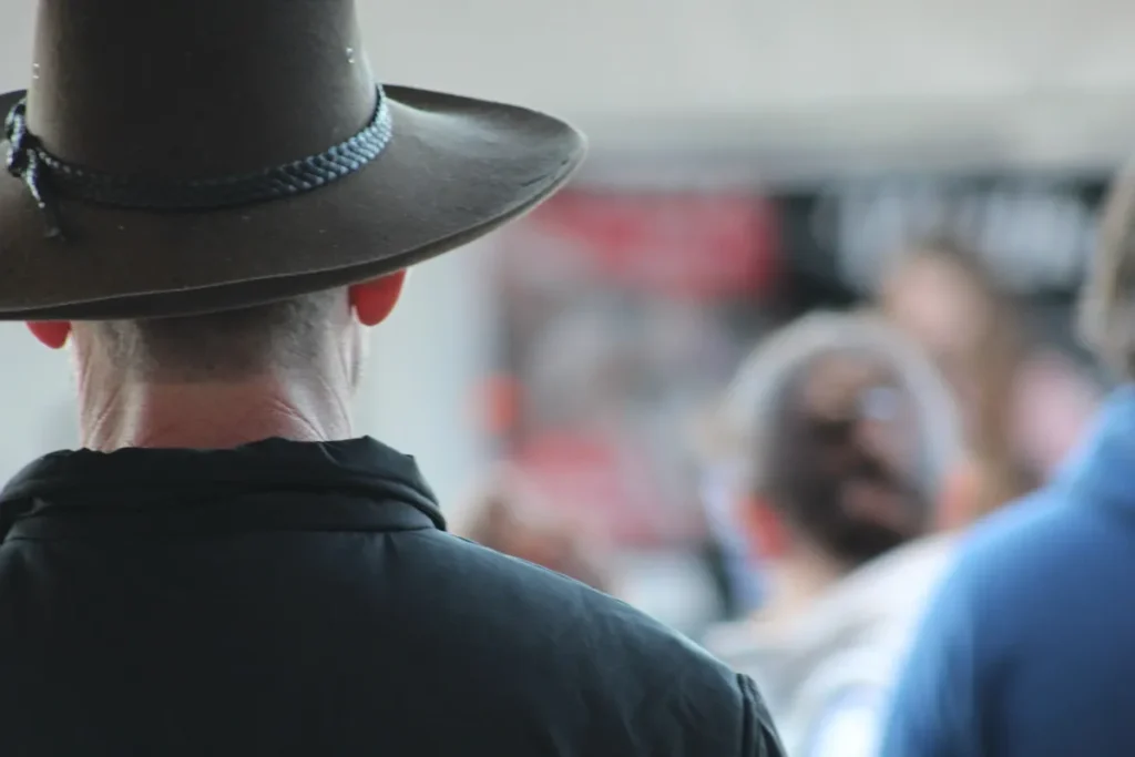 Back view of a person wearing a wide-brimmed Australian outback hat in a public outdoor setting with people in soft focus in the background.