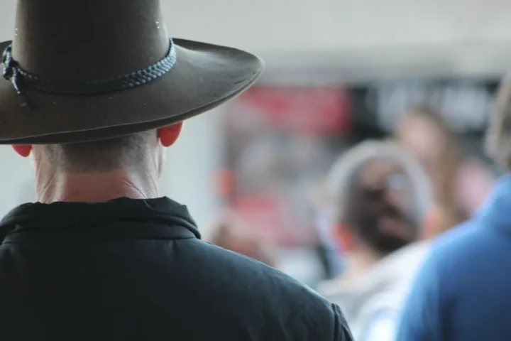 Back view of a person wearing a wide-brimmed Australian outback hat in a public outdoor setting with people in soft focus in the background.