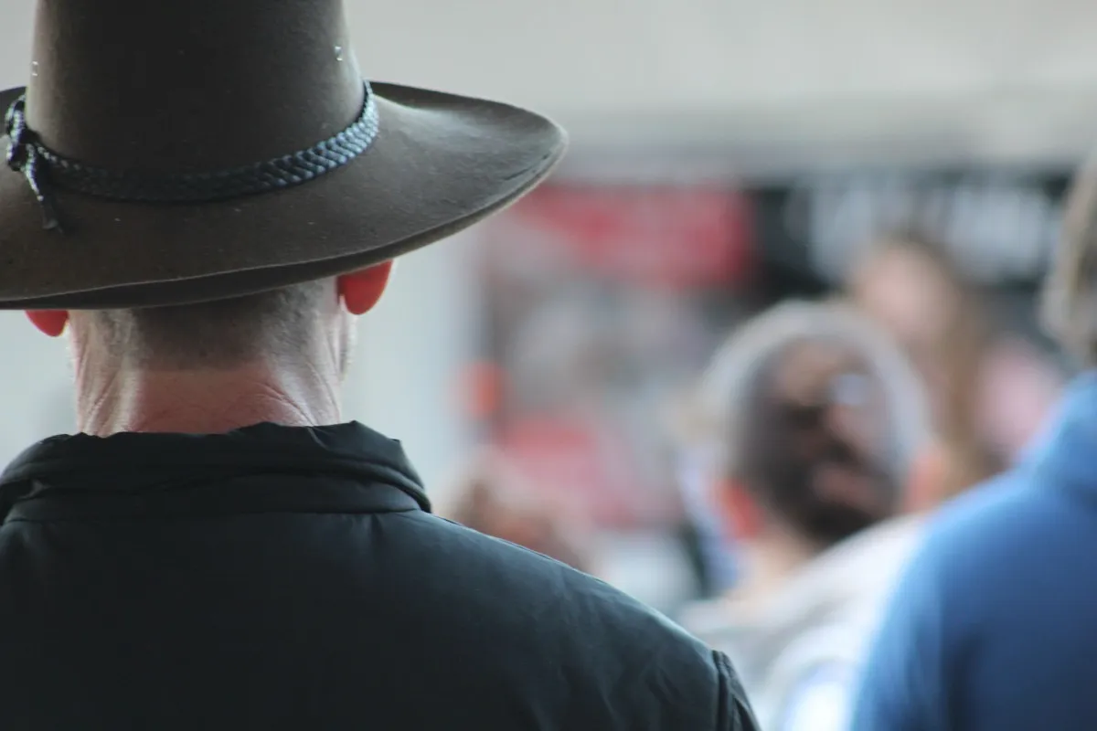 Back view of a person wearing a wide-brimmed Australian outback hat in a public outdoor setting with people in soft focus in the background.
