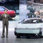 Toyota executive presenting a futuristic Corolla electric concept vehicle on stage with historic Corolla models displayed in the background.