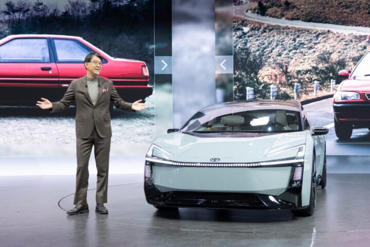 Toyota executive presenting a futuristic Corolla electric concept vehicle on stage with historic Corolla models displayed in the background.