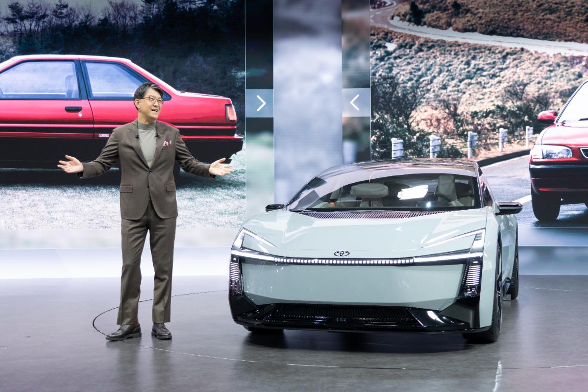 Toyota executive presenting a futuristic Corolla electric concept vehicle on stage with historic Corolla models displayed in the background.