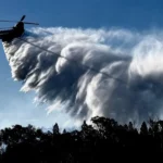 Helicopter operated by NSW RFS releasing a heavy water drop over forested terrain during bushfire operations in New South Wales on December 1, 2025.