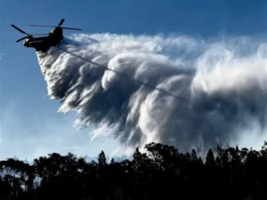 Helicopter operated by NSW RFS releasing a heavy water drop over forested terrain during bushfire operations in New South Wales on December 1, 2025.