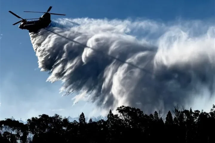 Helicopter operated by NSW RFS releasing a heavy water drop over forested terrain during bushfire operations in New South Wales on December 1, 2025.