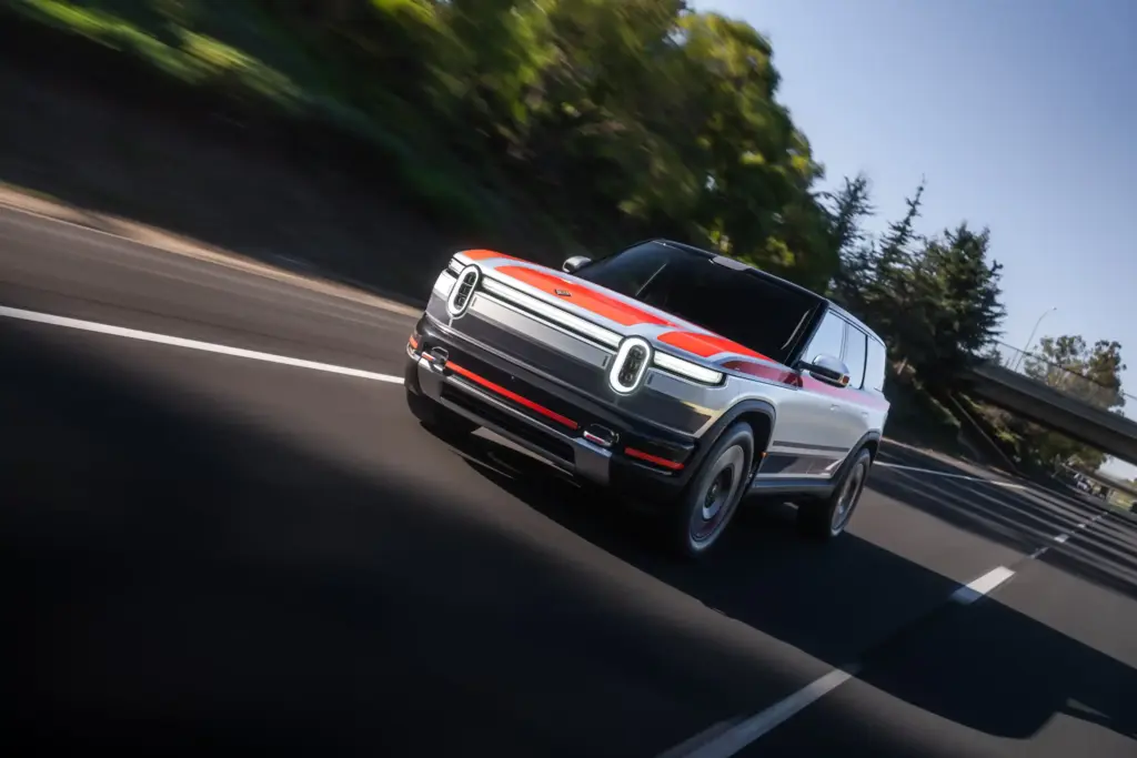 2026 Rivian R2 electric SUV prototype driving on highway during testing with orange and silver exterior and signature vertical LED headlights
