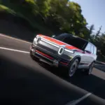 2026 Rivian R2 electric SUV prototype driving on highway during testing with orange and silver exterior and signature vertical LED headlights