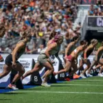 Female athletes sprinting from the start line during a CrossFit Games competition in a packed stadium.