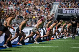Female athletes sprinting from the start line during a CrossFit Games competition in a packed stadium.